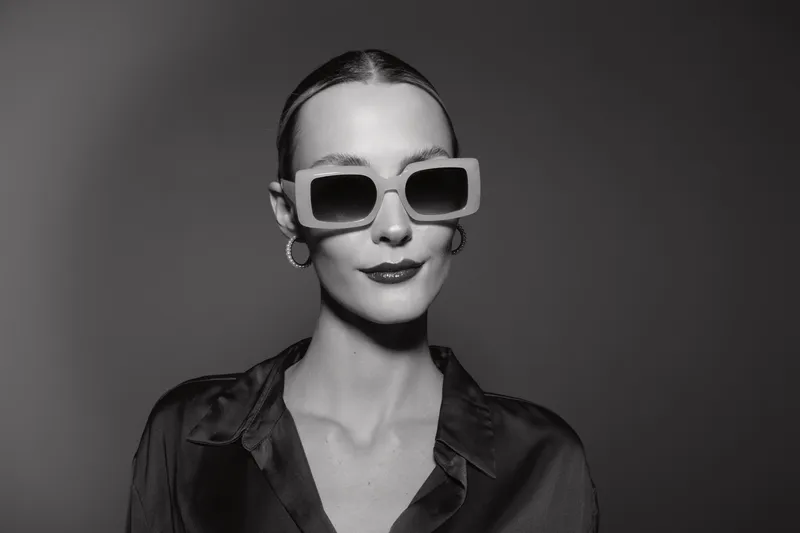 Young white woman with pale skin and dark hair wears oversized sunglasses and a silk blouse. Studio portrait with minimalistic style and confident expression.