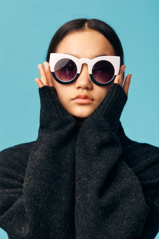 Young woman wearing oversized white sunglasses and black sweater posing against blue background. Fashion portrait with stylish glasses, casual outfit and confident expression in studio.
