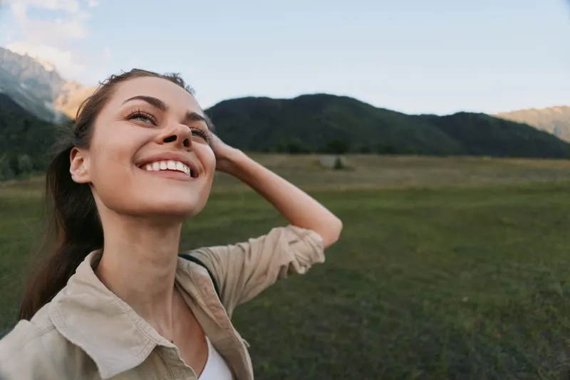 Young woman with long brown hair smiles joyfully while standing in a green meadow with mountains in the background under a clear sky.