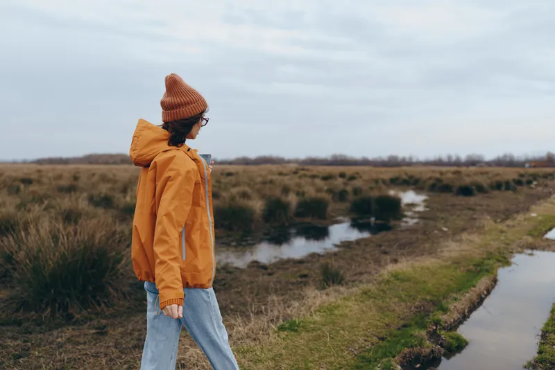 Woman lifestyle outdoors walking on countryside path, wearing orange jacket and knit hat, jeans, calm expression, standing near stream, cloudy sky background, natural light, peaceful