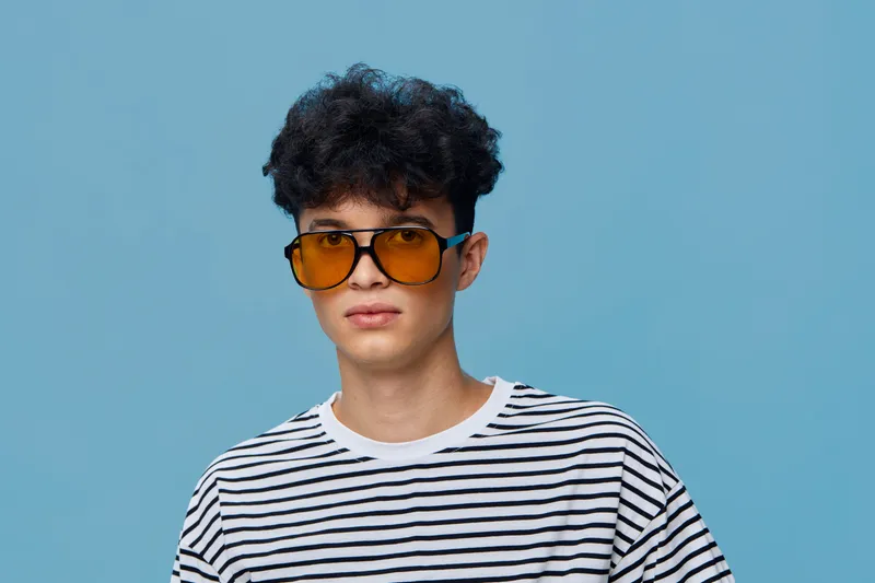 young man wearing orange sunglasses and striped shirt against blue background, casual style, curly hair, studio portrait with trendy vibe.