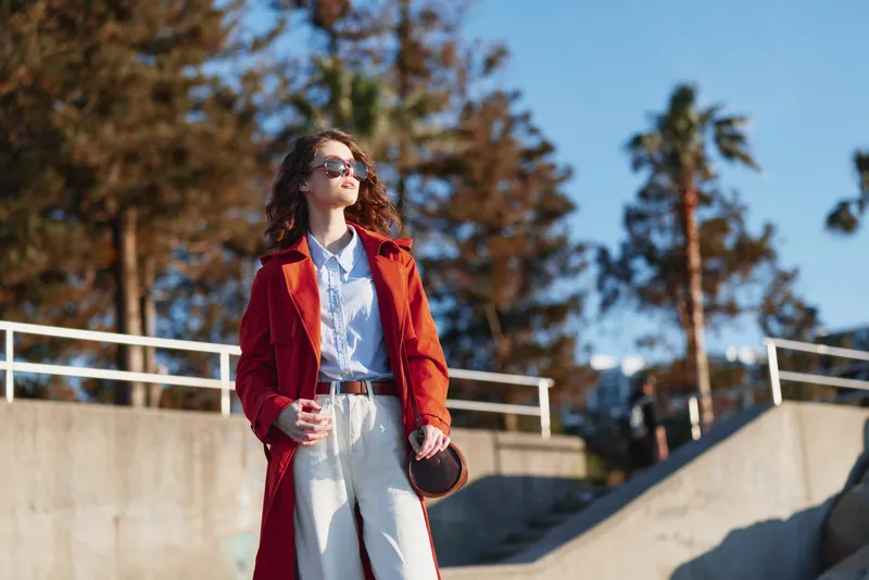 Candid woman outdoors wearing a red coat, sunny day, relaxed posture, soft breeze, natural colors, railing backdrop, fashion forward mood, confident expression