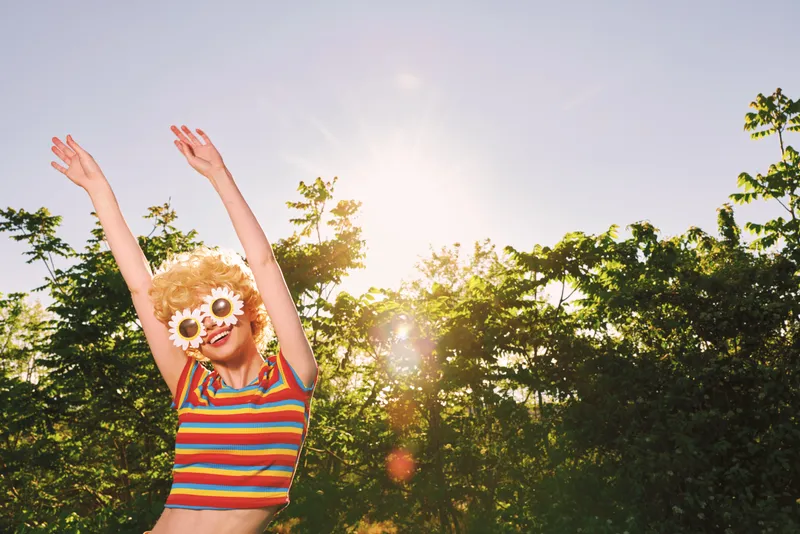 Cheerful woman outdoors, striped top, flower sunglasses, arms raised, sunlit smile, carefree lifestyle moment, warm retro tones, lush greenery background, candid playful energy
