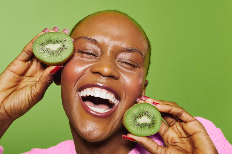 Happy African woman with dark skin holding two halves of fresh kiwi fruit near her face on green background. Concept of healthy eating, vitamin C, wellness, and natural beauty care.