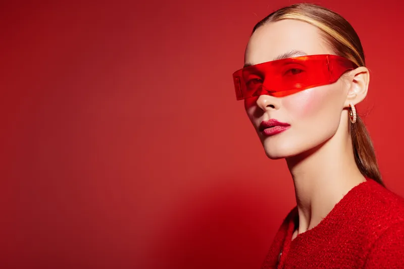 Stylish woman with red translucent sunglasses and sleek hair poses on red background. Fashion portrait with bold makeup and modern accessories. Concept of style and confidence.