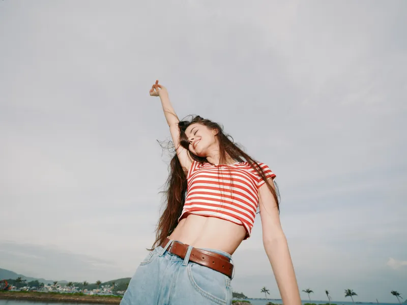 Vibrant young woman dances outdoors, lifting her arm in a carefree pose under a clear sky, wearing a red striped crop top and denim jeans, radiating freedom and joy.