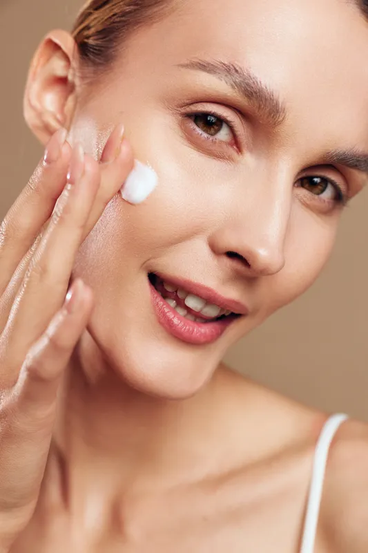 Young woman with brunette hair applies moisturizer to her face, smiling softly as she pats on cream on her cheek in a clean studio setting, conveying skincare, self care, and healthy glowing skin.