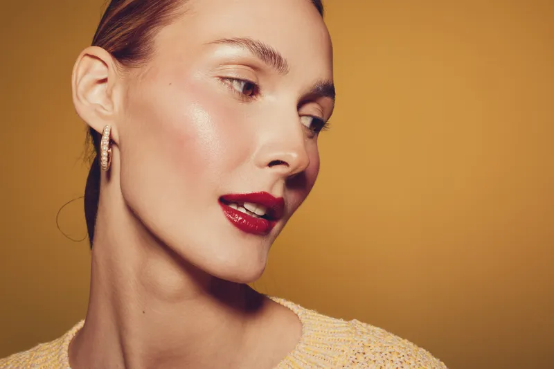 Close-up of a young woman with glowing skin and red lipstick looking sideways on a warm brown background. Concept of beauty, makeup, skincare, and fashion portrait.