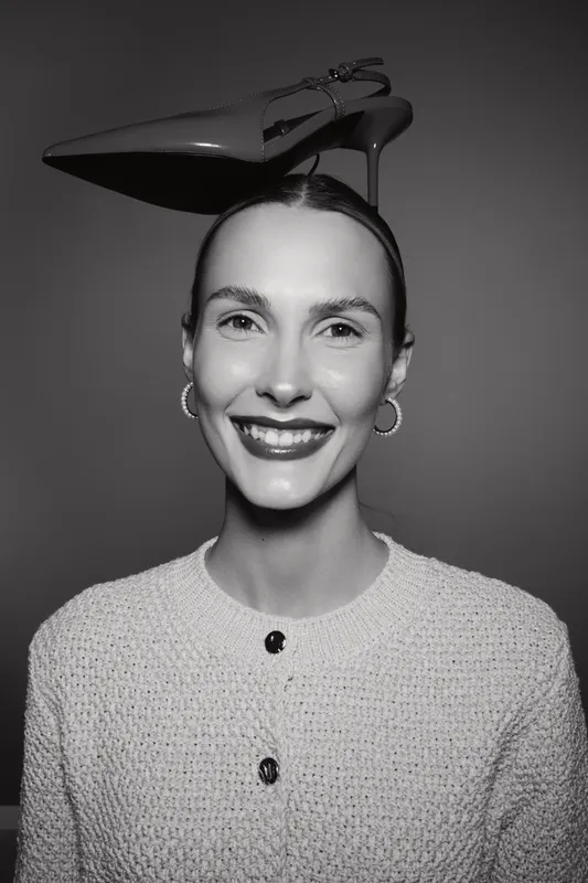 Smiling caucasian woman with pale skin and dark hair wears high heel shoe on head. Studio portrait in black and white with cozy sweater and hoop earrings.