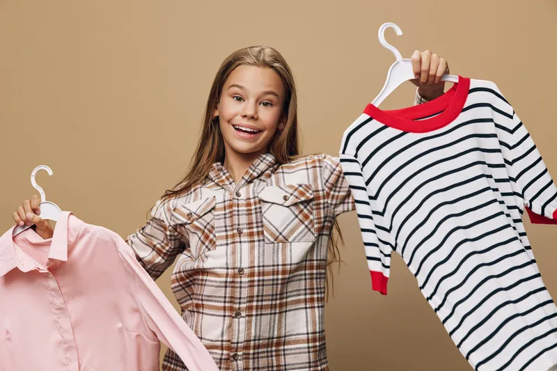 Girl holding two shirts on hangers smiling with casual plaid shirt and isolated brown background for clothing choice and fashion shopping style.