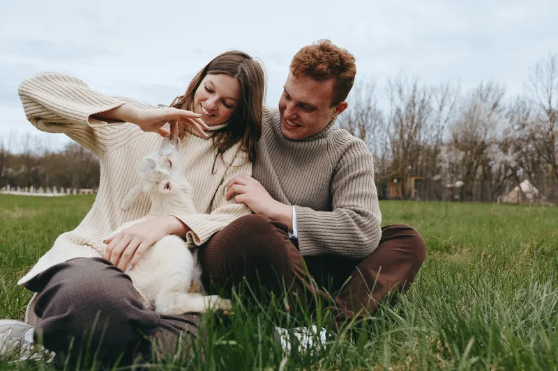 Couple outdoors sharing gentle affection with a playful goat kid in a sunlit meadow, cozy sweaters, relaxed posture, natural colors, candid moment of connection and joy