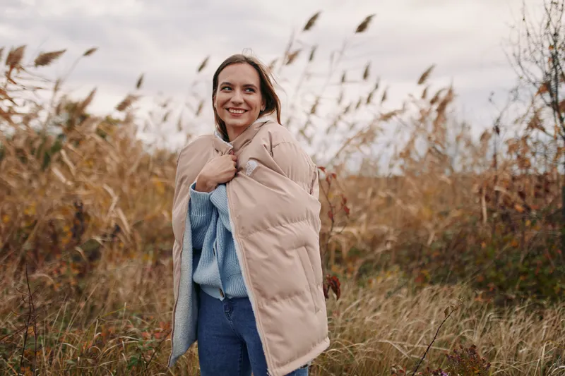 An authentic portrait of a smiling woman outdoors in autumn light, wearing a beige puffer and cozy sweater, conveying authenticity and accuracy of the moment.