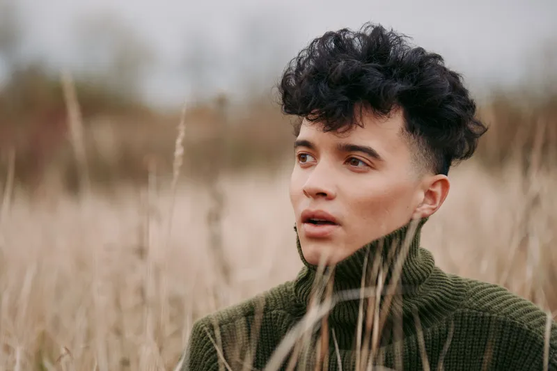 A candid close portrait of a young adult in a textured turtleneck amidst tall dry grass, focused gaze capturing an authentic expression, realistic skin tones and accurate emotional detail.