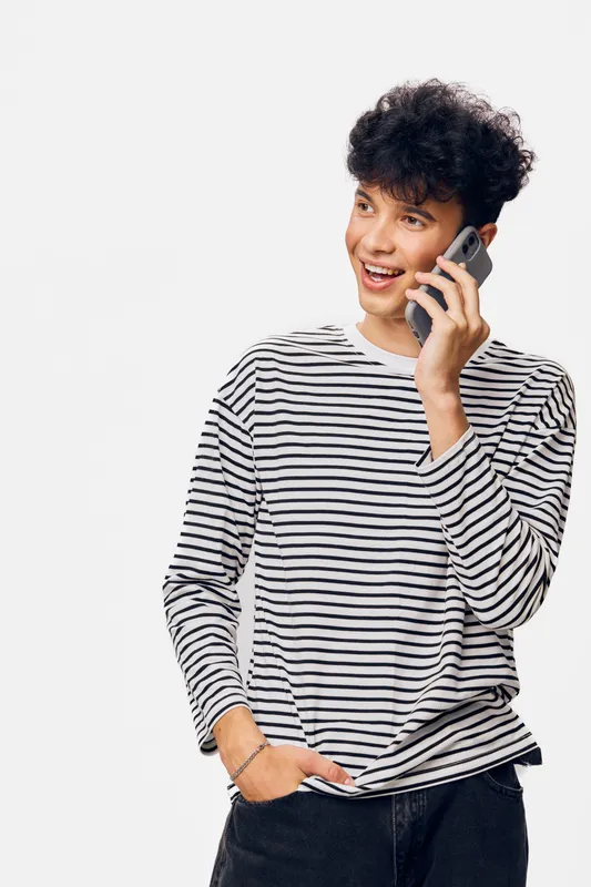 young man talking on smartphone smiling, wearing striped long-sleeve shirt and black pants, casual style with curly hair, isolated white background