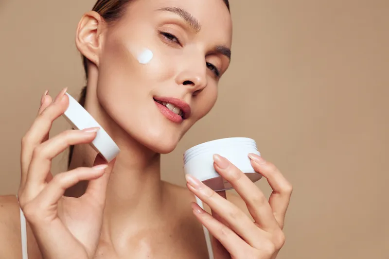 Young woman with clear skin applies moisturizing cream to her face while holding a white jar against a neutral beige background.