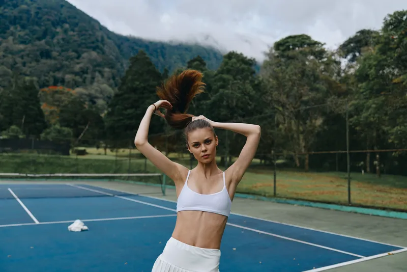 Young woman in sporty outfit stretching her ponytail on tennis court surrounded by nature, conveying focus and fitness vibes