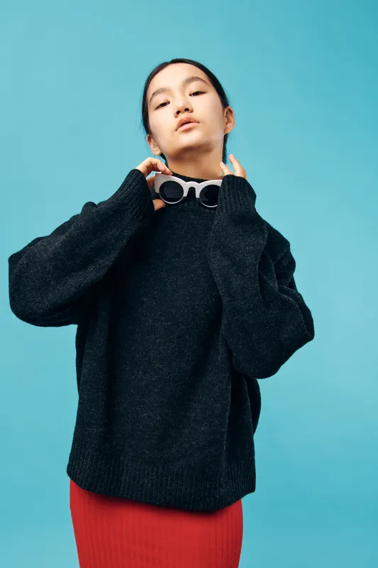 Young woman wearing black sweater and red skirt holding sunglasses on blue background. Stylish casual outfit with confident expression and minimal makeup in studio portrait.