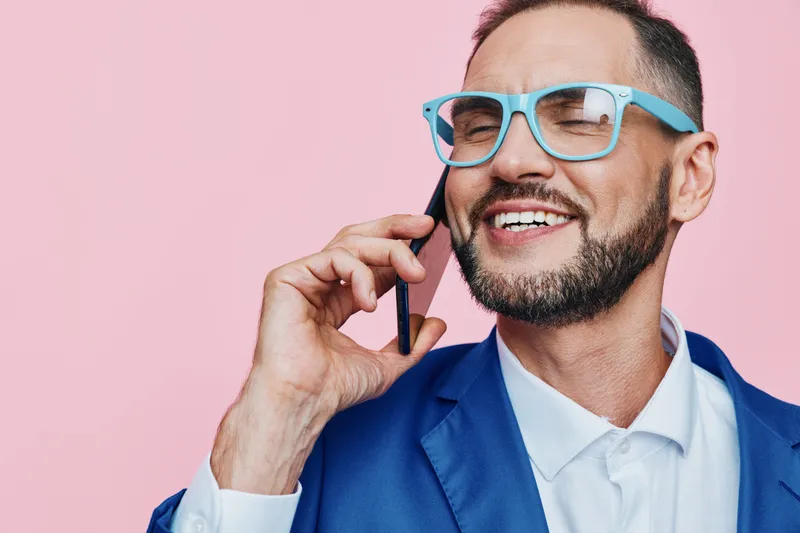 A professional man on a pink background chats on a mobile phone, radiating confidence and happiness, presenting a polished business demeanor suited for client engagement and teamwork in a modern