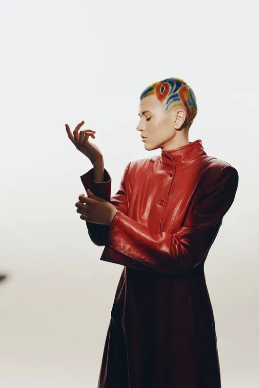 Woman with colorful short hair wearing a red leather jacket posing with delicate hand gestures, showcasing modern fashion and artistic expression in a minimal studio background.