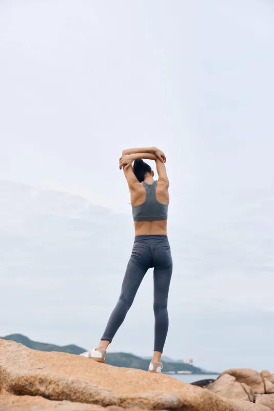 Female fitness model stretching on rocks by the sea, wearing sporty gray clothing, embracing nature and fitness for a healthy lifestyle