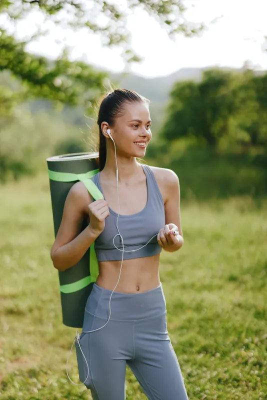 Young woman in gray activewear smiling while carrying a yoga mat outdoors, embodying fitness and wellness in a serene green environment