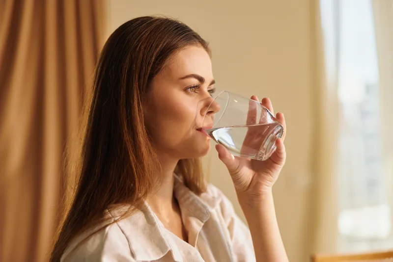 Woman drinking water from a glass at home, portrait profile in natural sunlight. Casual shirt and long hair, hydration moment for healthy lifestyle and daily wellness by the window.