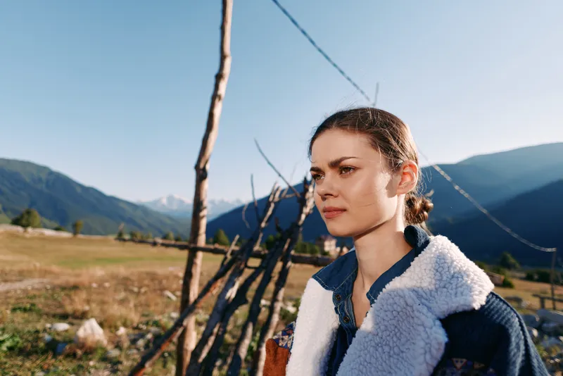 Woman portrait in countryside mountains, rural outdoors with wooden fence and sunlight, thoughtful female gaze wearing warm jacket and coat, serene landscape and natural beauty.