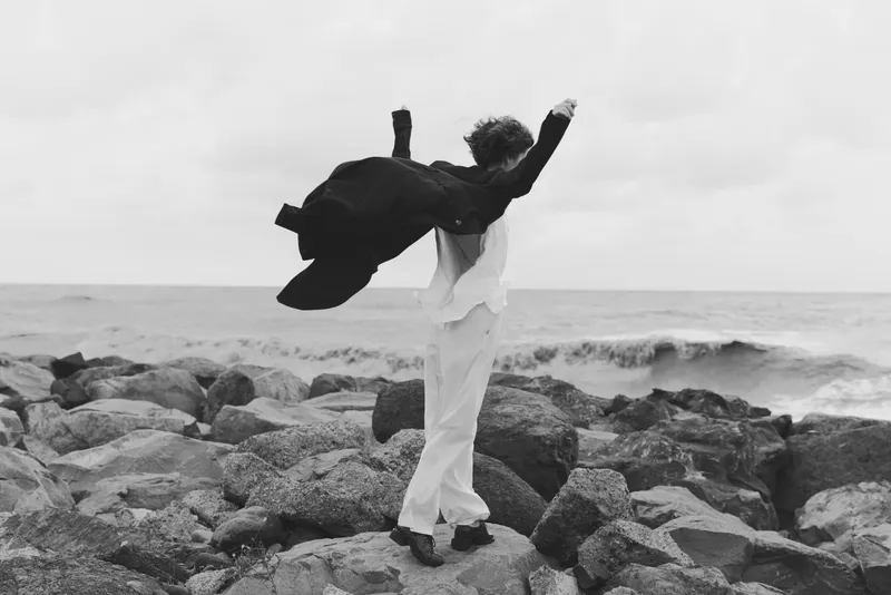White suit on rocky shoreline, jacket thrown high in motion as waves crash, black and white scene capturing freedom, celebration, motion