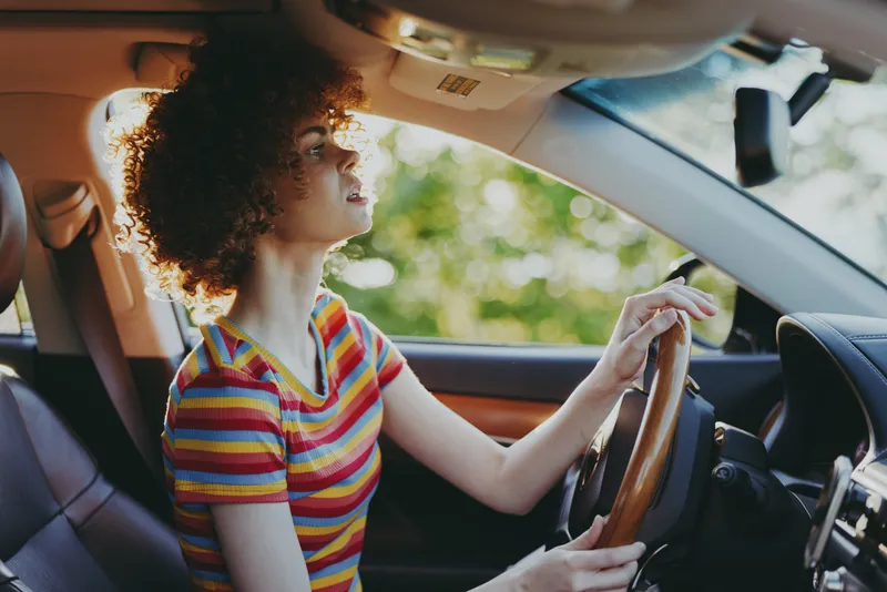 A woman driver sits in a car interior, striped shirt, curly hair, and a focused expression. Bright daytime scene suggests lifestyle, travel, and retro film color.