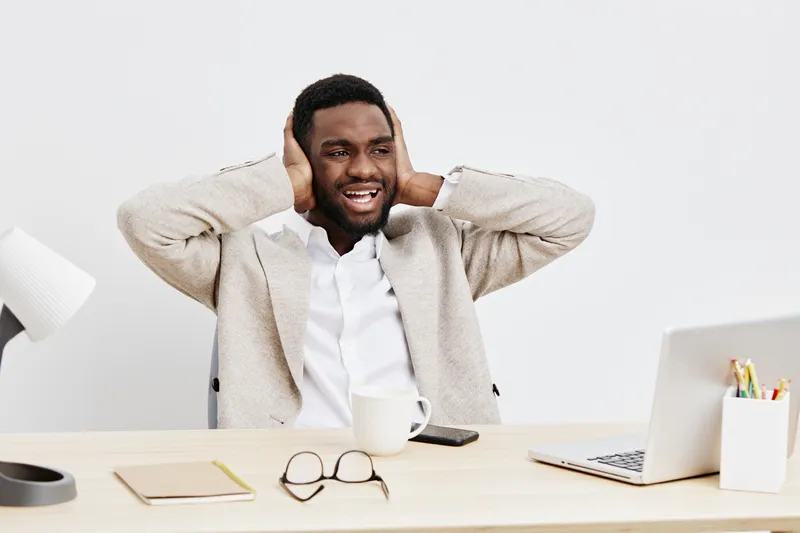 Smiling man with dark curly hair and beard, wearing beige cardigan and white shirt, sitting at desk with laptop, coffee cup, glasses, and notebook, expressing happiness and relaxation in a bright mode