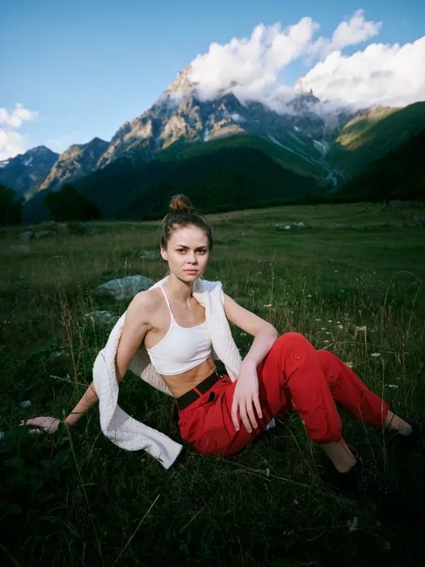 Woman in white top and red pants sits in a meadow with towering mountains and clouds, outdoor fashion and nature inspiration for travel, leisure, and adventure photography