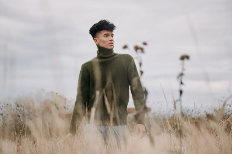 An authentic portrait of a young man standing in tall grass, wearing a dark green turtleneck, conveying a calm, grounded presence in a wide field.