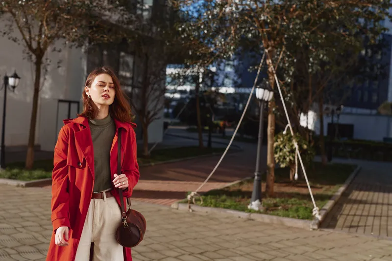 Young woman in a red coat walks confidently along a sunny urban street holding a round handbag, with autumn trees and modern buildings in the background.