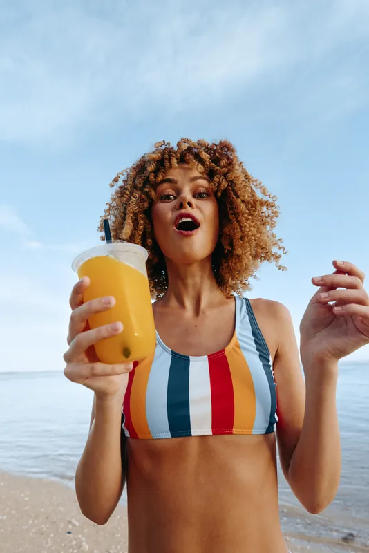 Young woman with curly hair holds a refreshing orange juice at the beach, wearing a striped bikini and smiling toward the camera.