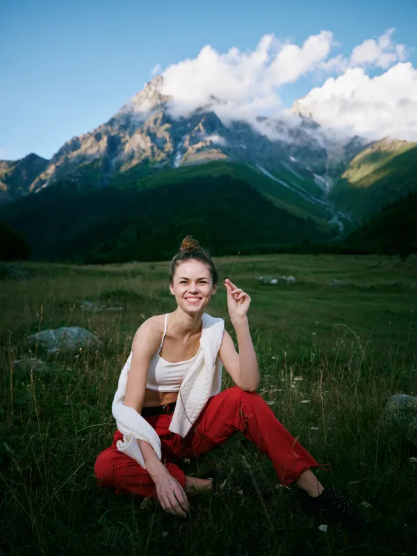 Smiling woman in red pants sits on a grassy meadow with mountains and blue sky behind, relaxed outdoor scene of nature, travel and adventure in a scenic alpine landscape