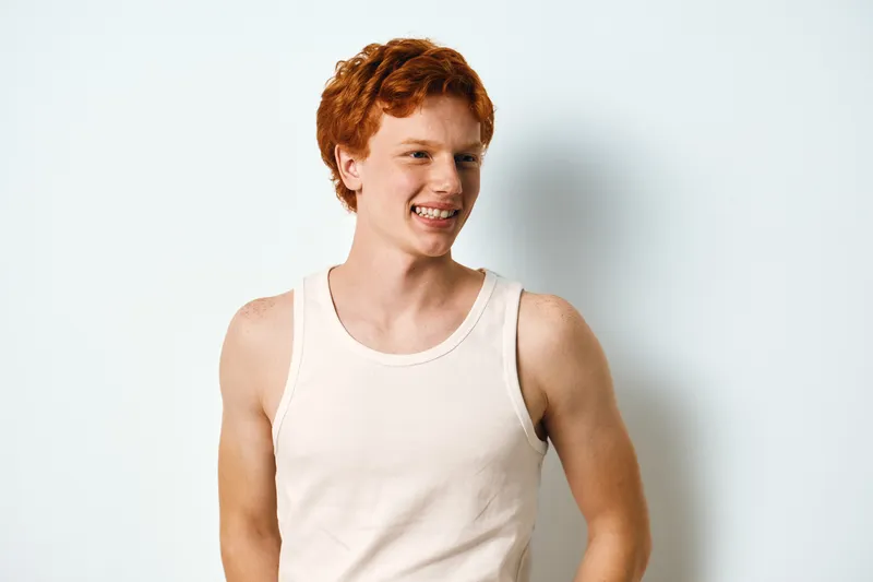 Young man with red hair and freckles wearing white tank top smiling and looking away on white background. Casual portrait, happy expression, isolated, studio shot with natural light.