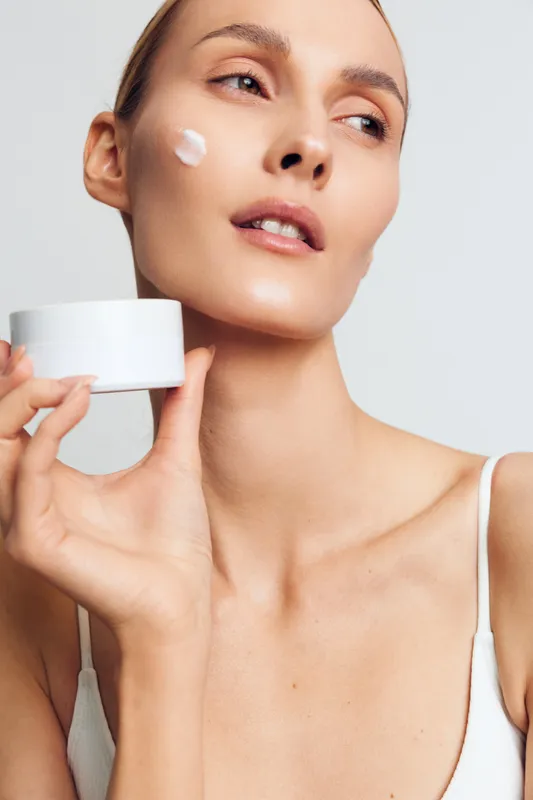 Young woman applying face cream on cheek, holding white cosmetic jar, clean skin, natural makeup, minimalistic background, skincare routine, healthy lifestyle concept