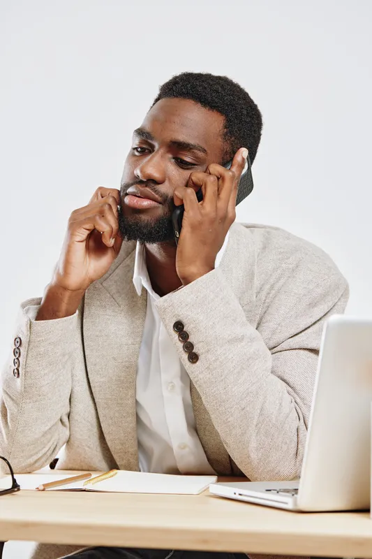Professional young african-american man in business casual attire speaking on smartphone while sitting at desk with laptop and notepad, indoors background, studio shot, natural lighting, candid busine