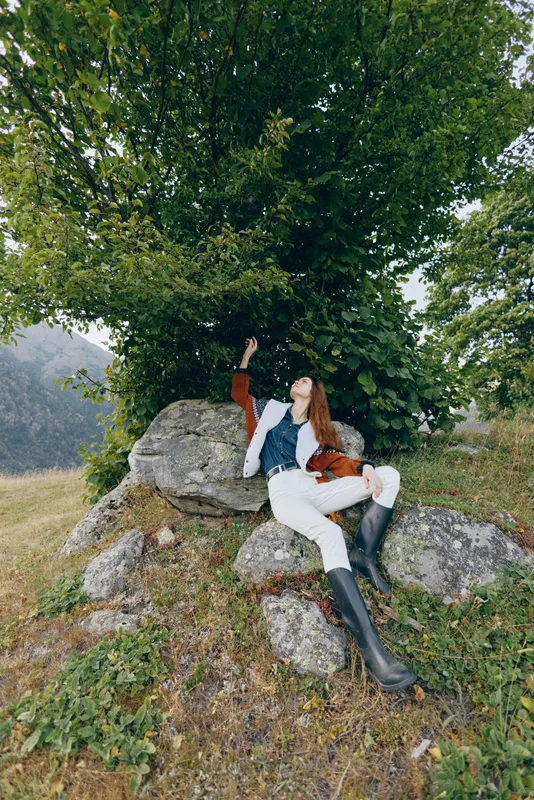 Woman relaxing on rocks under a tree in the countryside nature, wearing boots and a casual jacket, enjoying outdoor serenity on a grassy hillside near mountain scenery.