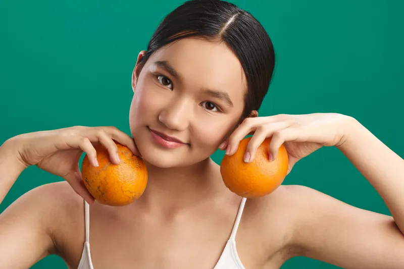 Asian woman smiles gently as she holds two oranges near her cheeks, posing playfully in a bright studio with a clean, vibrant background against a solid backdrop.
