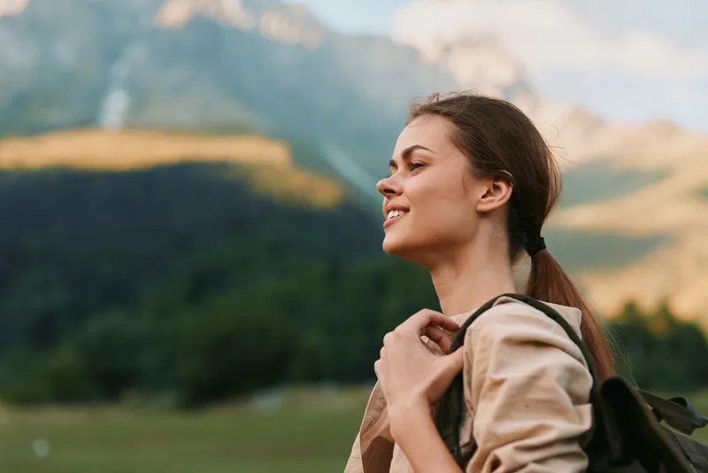 A woman in outdoor gear enjoys a sunny hike, backpack secured, as rugged mountains rise in the background. A calm moment for wellness, exploration, and everyday adventure.