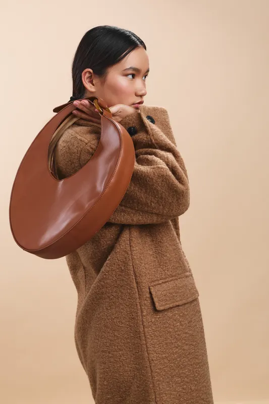 asian woman wearing a woolen coat carries a stylish brown bag over her shoulder in a minimalist studio setting, exuding poise and modern fashion.