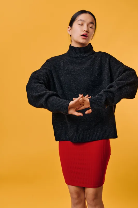 Young woman in black sweater and red skirt standing with eyes closed and hands clasped in front of yellow background for fashion and style concept.