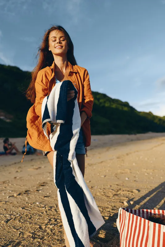 Young woman on sandy beach holding striped towel, smiling with closed eyes, wearing orange shirt, sunny day, green hills in background, relaxed mood