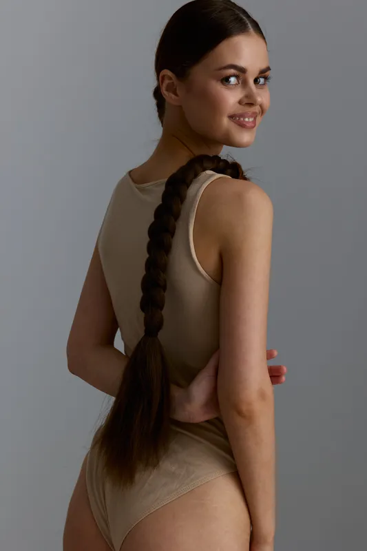 smiling woman with long braid wearing beige bodysuit looking over shoulder against gray background, natural light, minimalist style