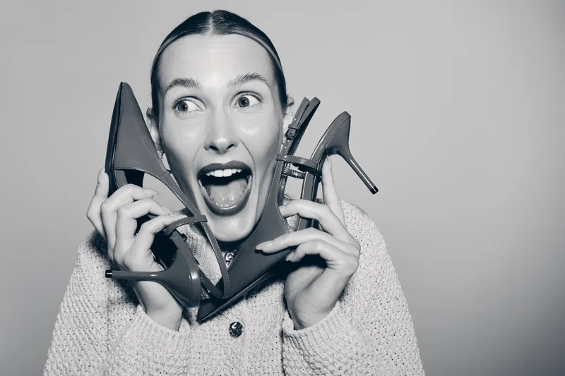 Young Caucasian woman with light skin holds two red high heels near her face, smiling with excitement against plain background. Concept of fashion, style, and happiness.