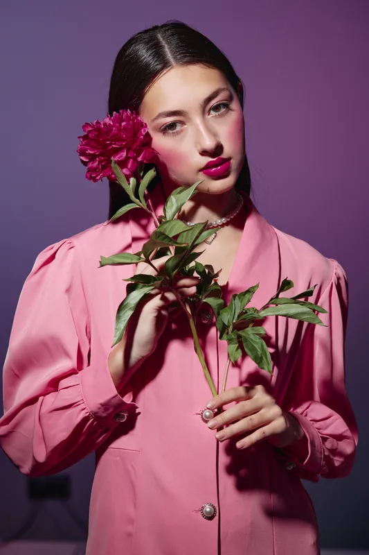 Young woman with striking dark hair holds a red peony against a lavender background. She wears a pink vintage-style dress. Creative beauty and fashion concept.