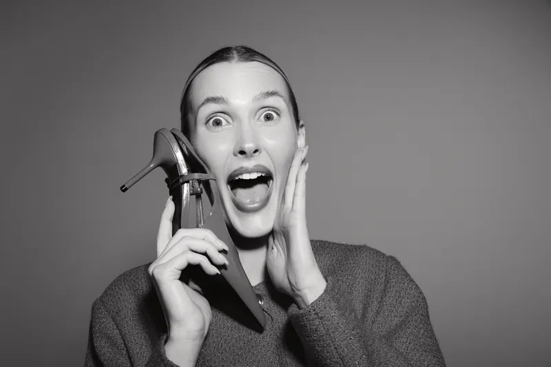Excited white woman with pale skin holds high heel shoe near face, expressing joy and surprise. Black and white studio portrait with emotional concept and fashion element.