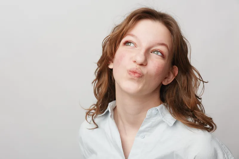 Young woman with curly hair wearing white shirt looks up thoughtfully on light background. Concept of contemplation, decision making, and casual lifestyle portrait.
