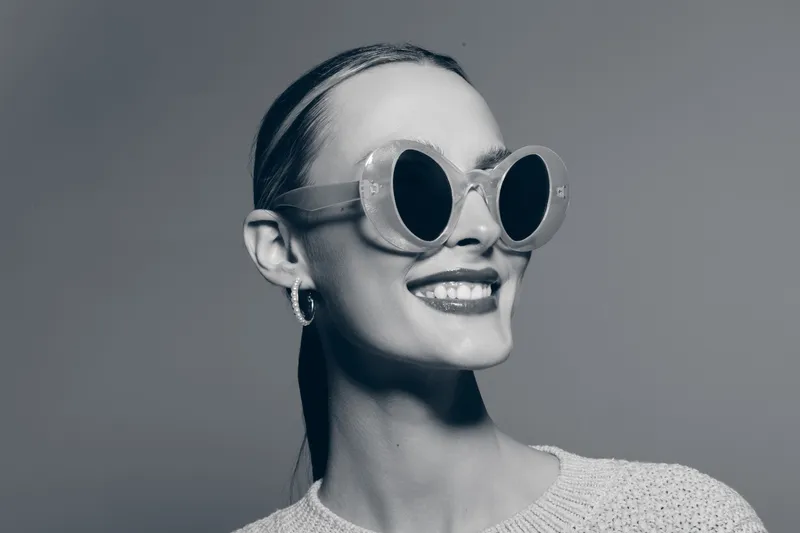 Portrait of a smiling Caucasian woman with light skin wearing round sunglasses and hoop earrings. Minimalist studio grey background emphasizes her stylish and confident look.